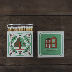 matchbox showing an oak tree on the front with acorns in each corner. The reverse of the box shows a victorian red brick building