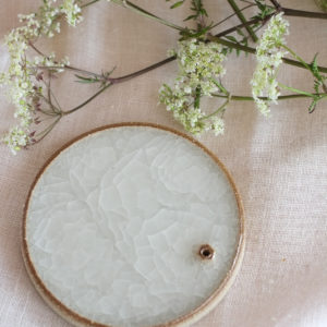 round, flat object on a white cloth background. The object has a flat but textured surface and is off-white in colour. There is a sprig of greenery with white flowers laying alongside the object. .