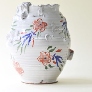 Round ceramic vessel shown on a pale cream background. The vessel is an off-white or very light grey in colour with hand painted flowers in blue and red.