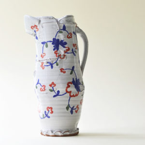 Pale white ceramic jug shown on a pale cream background. The jug has a wonky, reconfigured look to it with handpainted blue and red vines on it.
