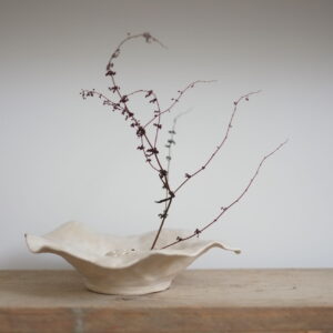 A shallow fluted vase made from an off-white stoneware. Shown on a wooden shelf against a white backdrop. There is a single stemp of bracken displayed in the vase.