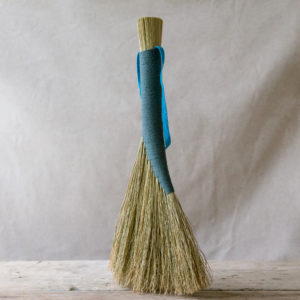 pale, natural coloured broom bound with a blue or teal coloured cord. Shown on a pale wooden table against a light fabric backdrop