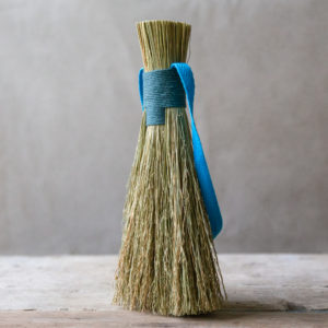 natural fibre hand brush, bound with a teal blue cord. Show standing on a pale wooded table against a light fabric backdrop.