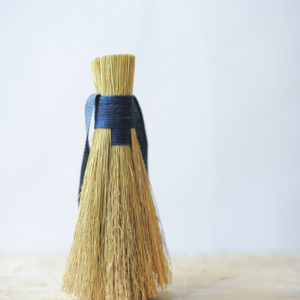 natural fibre hand brush, bound with a hand dyed indigo cord. Show standing on a pale wooded table against a white backdrop.