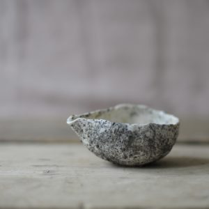 a small white and grey, heavily textured pouring vessel set against a pink linen backdrop, on a rustic wooden board.
