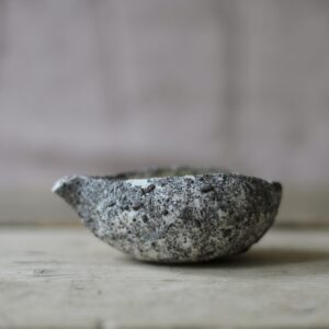 a small white and grey, heavily textured pouring vessel set against a pink linen backdrop, on a rustic wooden board.