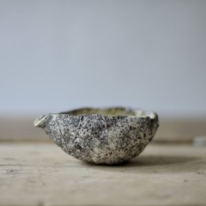 a small white and grey, heavily textured pouring vessel set against a pink linen backdrop, on a rustic wooden board.