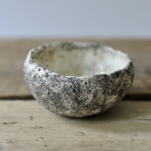 a small white and grey, heavily textured tea bowl set against a white backdrop, on a rustic wooden board.