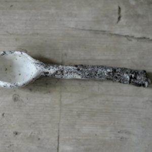 a small white and grey, heavily textured cawl spoon on a rustic wooden board.