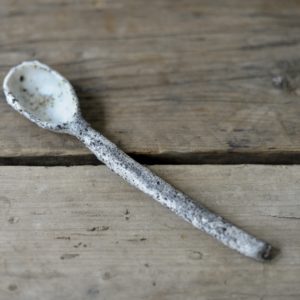 a white and grey, heavily textured cawl spoon on a rustic wooden board.