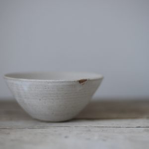 A cone shaped stoneware bowl with a pale matt finish, shown on a rustic wooden tabletop against a light grey background.