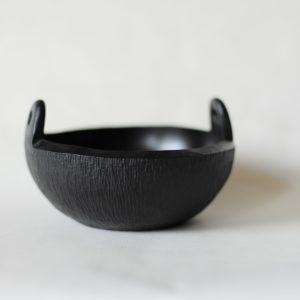 A black, hand carved bowl with small handles on either side. Shown against a white backdrop.