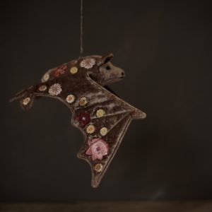A pinkish grey, velvet bat, embroidered with colourful flowers, shown in mid flight against a dark background.