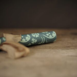 A bluey/green coloured wrapping paper with a repeat flower pattern. Photographed rolled and tied up in a bow with a light brownish pink, silk ribbon. Laying on a rustic wooden tabletop against a very dark background.