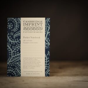 A dark/light blue paisley patterned paperback notebook with a Cambridge Imprint label on the front. Photographed on a rustic wooden table against a dark brown, almost black background.