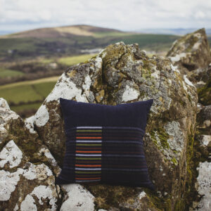 Dark blue cushion with multi-coloured stripes. Photographed against a craggy hillside out in the welsh landscape