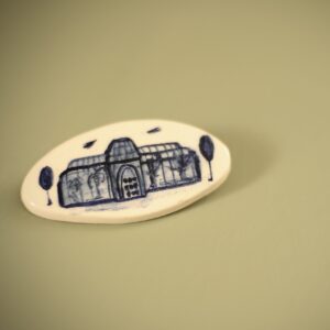 A white and blue ceramic brooch with a naive illustration of the a botanical glass house.